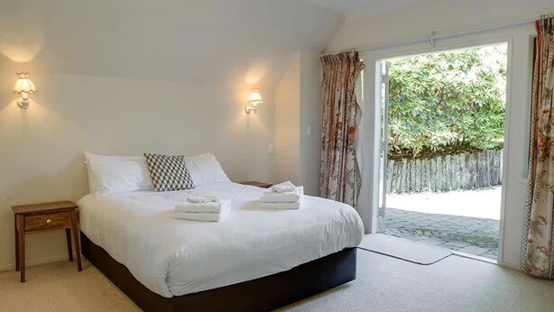 A bedroom with a large bed, two bedside lamps, a small nightstand, and a view of a garden through a sliding glass door. - Mountain Bike Manor - Spa Pool, Sleeps 8, Pets OK (Rotorua)