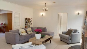 A living room with a grey sofa, a grey armchair, a coffee table, and a fireplace. There is a bookshelf and a chandelier. - Mountain Bike Manor - Spa Pool, Sleeps 8, Pets OK (Rotorua)