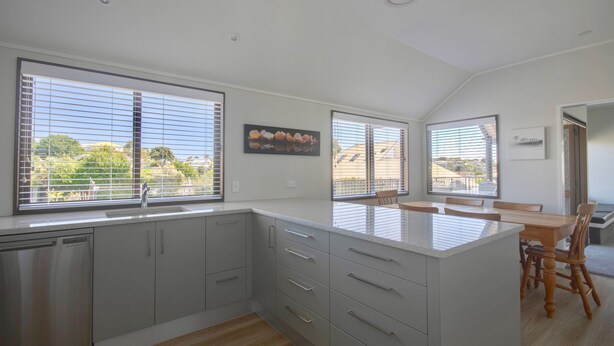 A modern kitchen with large windows, a central island, and a dining area with wooden chairs.
