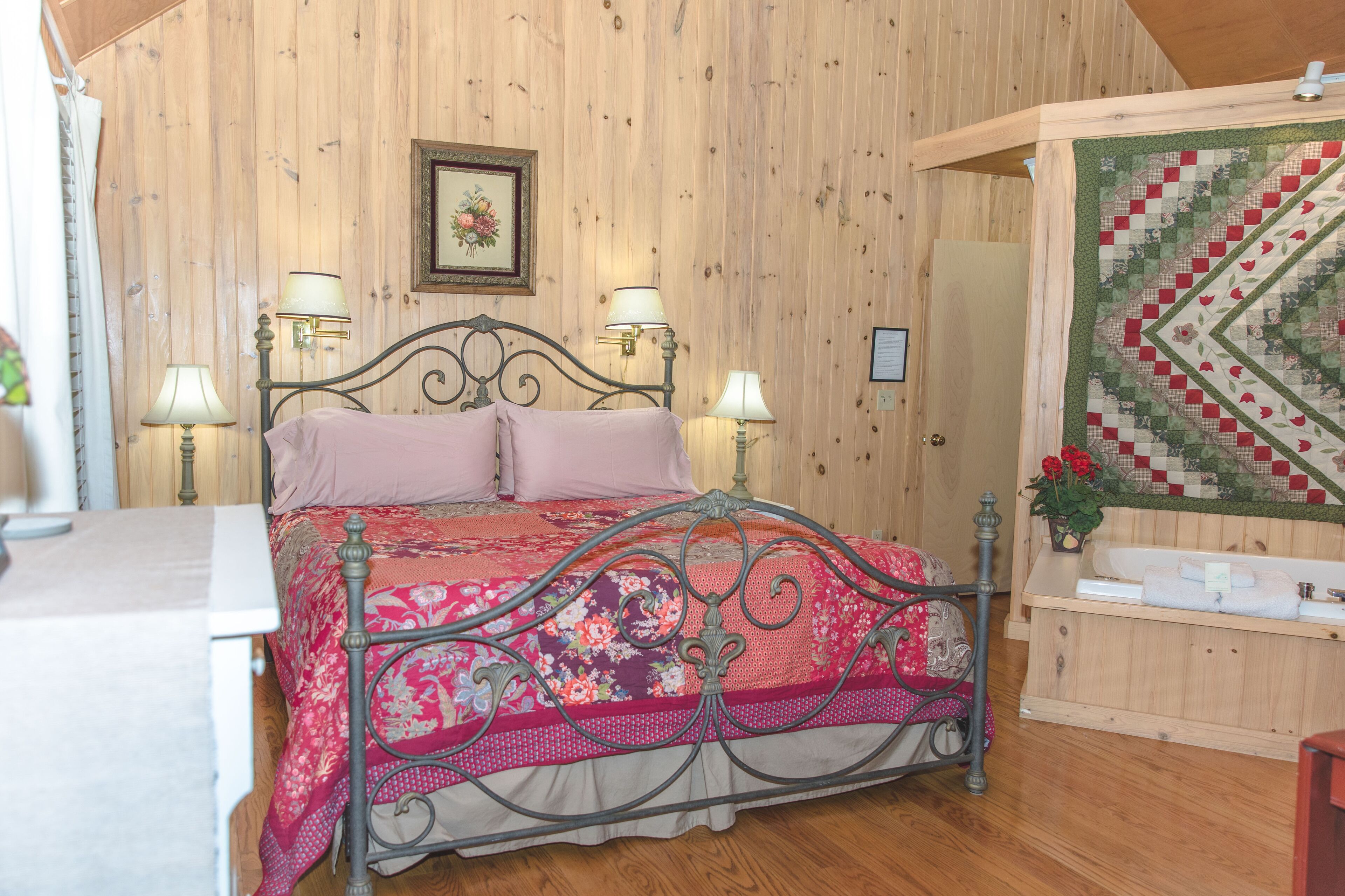 A wooden-paneled room with a bed, two wall-mounted lamps, a framed picture, and a quilted wall hanging.