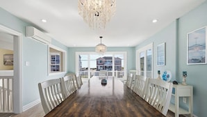 A spacious dining room with a long wooden table, white chairs, a chandelier, and a view of a house through large windows.
