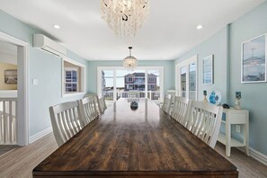 Uma sala de jantar espaçosa com uma longa mesa de madeira, cadeiras brancas, um candelabro e vista para uma casa através de grandes janelas.