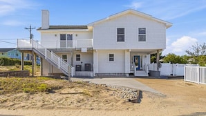 A two-story white house with a covered porch, a staircase, and a sandy beach in the background.