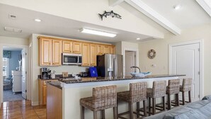 A kitchen with wooden cabinets, a stainless steel refrigerator, a microwave, and a bar area with wicker stools.