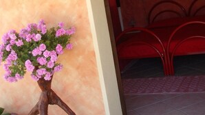 A room with a pink wall, a wooden stand with pink flowers, a red bed, and a painting on the wall. - Domu de Palla (Bauladu)