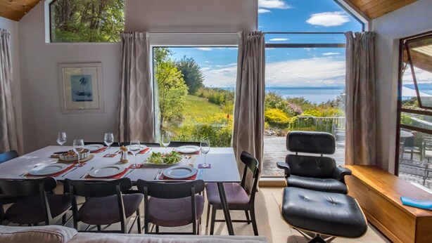 A dining area with a table set for six, large windows offering a view of greenery and the sea, and a cozy seating area with leather chairs.