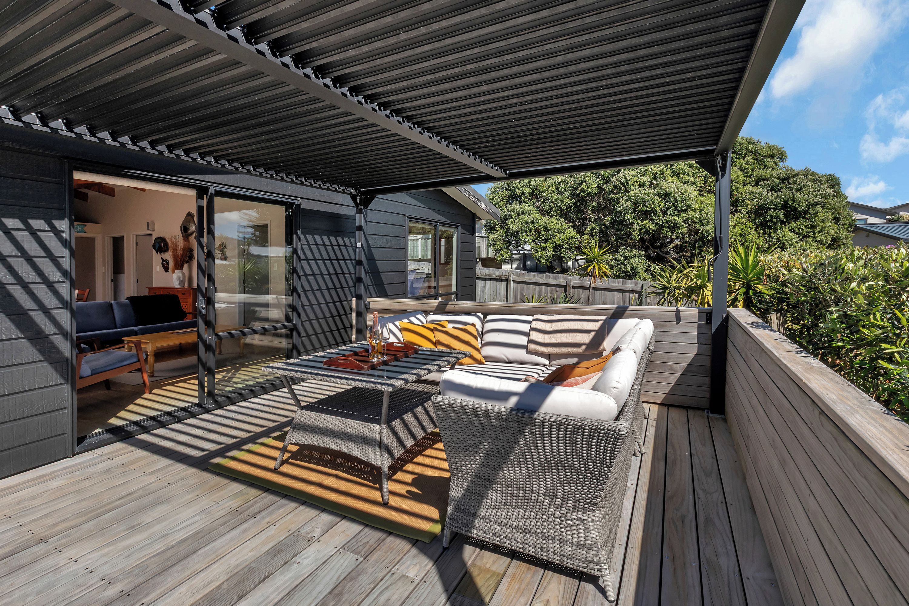 Un espace extérieur aménagé, abrité et doté d’un canapé et d’une table, ainsi que d’une terrasse en bois offrant une vue sur une maison et des arbres.