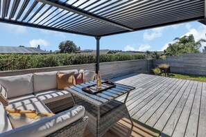 An outdoor patio with a wooden deck, a cushioned sofa with pillows, and a glass table with a tray and wine bottle. - The Hideaway - Omaha Holiday Home (Omaha)