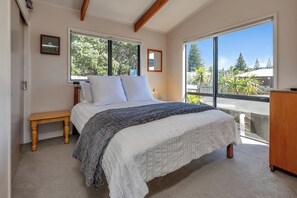 A bedroom with a bed, a bench, a desk, and large windows with a view of trees. - The Hideaway - Omaha Holiday Home (Omaha)
