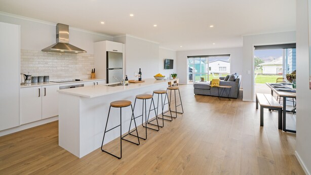 A modern kitchen with a central island, bar stools, and a dining area with a bench. - Seascape - Cooks Beach Holiday Home (Cooks Beach)