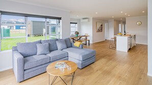 A modern living room with a grey sectional sofa, a round coffee table, and a kitchen area with a white countertop and bar stools. - Seascape - Cooks Beach Holiday Home (Cooks Beach)
