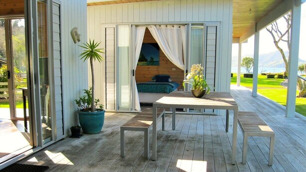 A wooden deck with a table and benches, a potted plant, and a sliding glass door leading to a room with a bed and curtains. - Paradise in Flaxmill Bay - Flaxmill Bay Home (Cooks Beach)