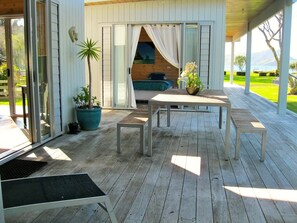 A wooden deck with a table and benches, a potted plant, and a sliding glass door leading to a room with a bed and curtains. - Paradise in Flaxmill Bay - Flaxmill Bay Home (Cooks Beach)