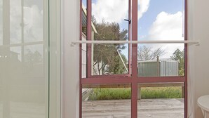 A room with a large window, a wooden deck outside, and a white wall. - Carefree at Cooks - Cooks Beach Bach (Cooks Beach)