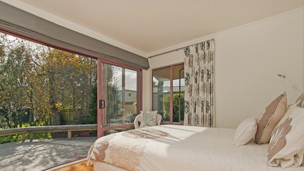 A bedroom with a large bed, a view of a balcony, and a window with curtains. - Carefree at Cooks - Cooks Beach Bach (Cooks Beach)