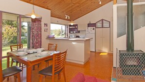 A modern kitchen with wooden ceiling beams, a dining area with a table and chairs, and a large window overlooking a garden. - Carefree at Cooks - Cooks Beach Bach (Cooks Beach)