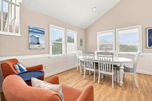 A dining area with a white table and chairs, a blue cushioned chair, and a painting of a beach house on the wall.