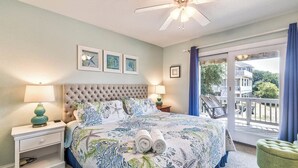 A bedroom with a tufted headboard, a large bed with a floral bedspread, two bedside lamps, a nightstand, a green ottoman, and a balcony with a view of a house and trees. - Ocean Paradise | 670 ft from the Beach | Private Pool, Hot Tub | Corolla (Corolla)
