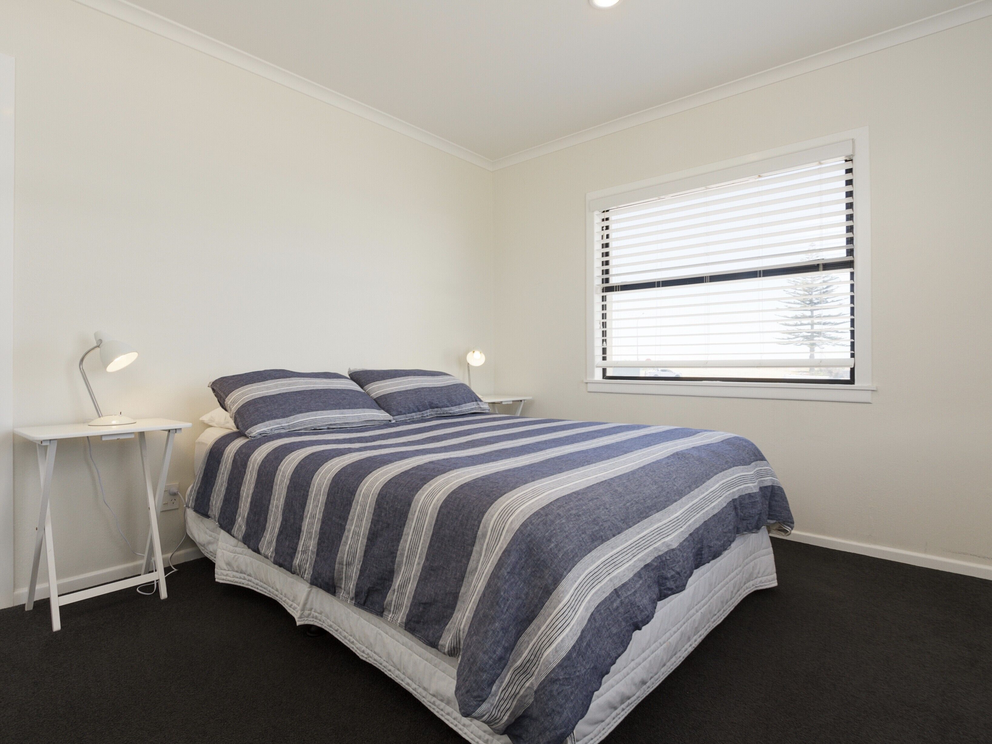 A bedroom with a bed, a bedside table with a lamp, a window with blinds, and a carpeted floor.