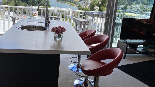 A modern kitchen with a view of a lake and mountains, a dining area with red chairs, and a TV. - Rooftop garden, Lakeview, 5 mins walk to town (Queenstown Hill)