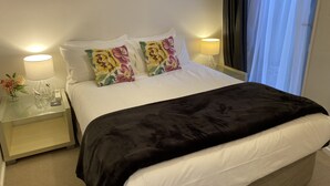 A neatly made bed with a black blanket, two floral pillows, and a white duvet. There are two bedside tables with lamps and a vase of flowers. - Rooftop garden, Lakeview, 5 mins walk to town (Queenstown Hill)