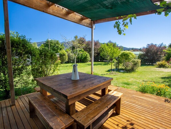 Una terraza de madera con una mesa de picnic y bancos, rodeada de abundante vegetación y una vista despejada del paisaje.