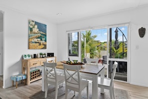A dining area with a table, chairs, a painting, and a view through large glass doors.