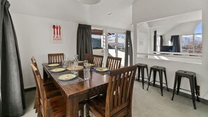 Un comedor con una mesa de madera preparada para cuatro personas, un televisor a la derecha y vistas a montañas a través del ventanal.