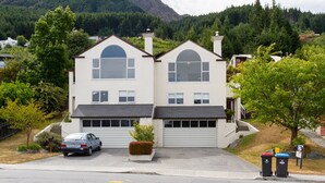 Una casa de dos pisos con garaje, rodeada de árboles y una montaña al fondo.