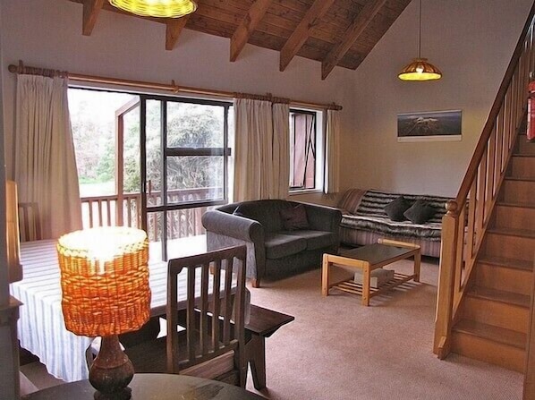 A cozy living room with a sofa, a table, and a lamp. There is a window with curtains and a view of trees outside.