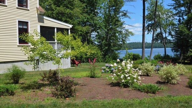 A house with a covered porch, a garden with various plants and flowers, and a lake in the background.
