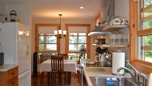 A kitchen with wooden cabinets, a refrigerator, a dining table with chairs, a chandelier, and a window with blinds.