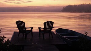 A wooden dock with two Adirondack chairs facing a calm lake at sunset.