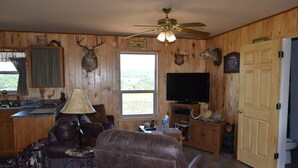 A cabin interior with a leather sofa, a ceiling fan, a mounted deer head, a television, and a wooden door. - Rustic Loyal Valley Private Guesthouse with Peace & Serenity Views (Mason)
