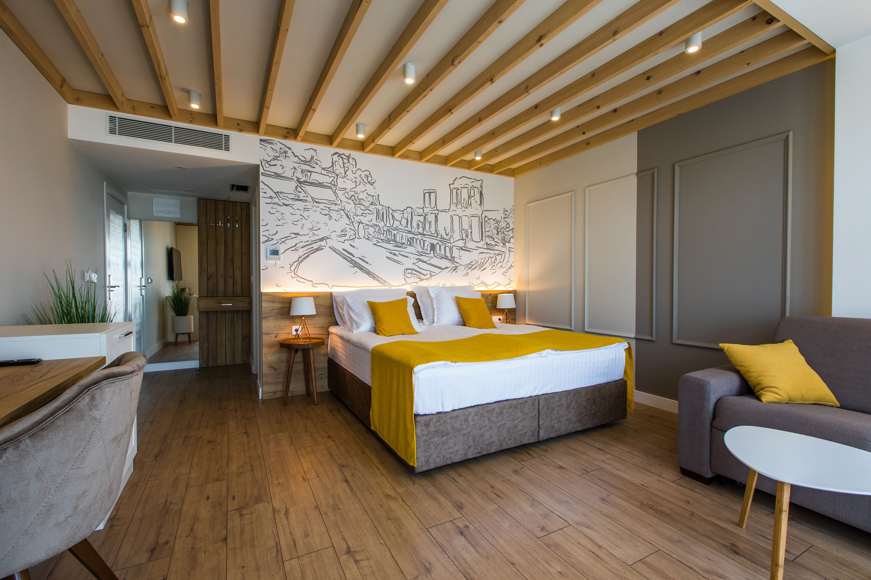 A modern hotel room with a wooden ceiling, a large bed with a yellow blanket, a grey sofa, and a white table. - HillHouse Plovdiv (Plovdiv)