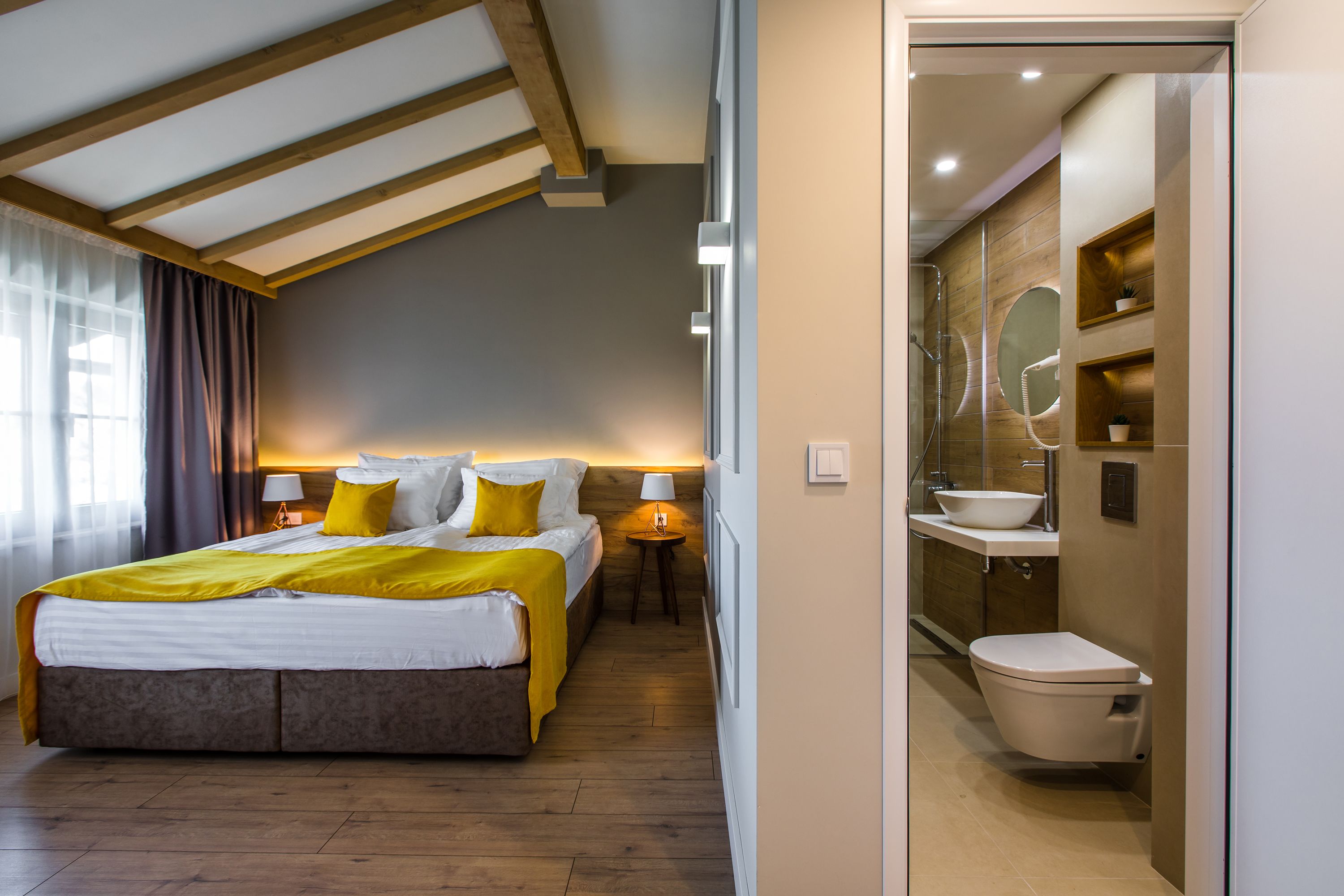 A modern hotel room with a large bed, bedside tables, a bathroom with a sink and toilet, and wooden beams on the ceiling.