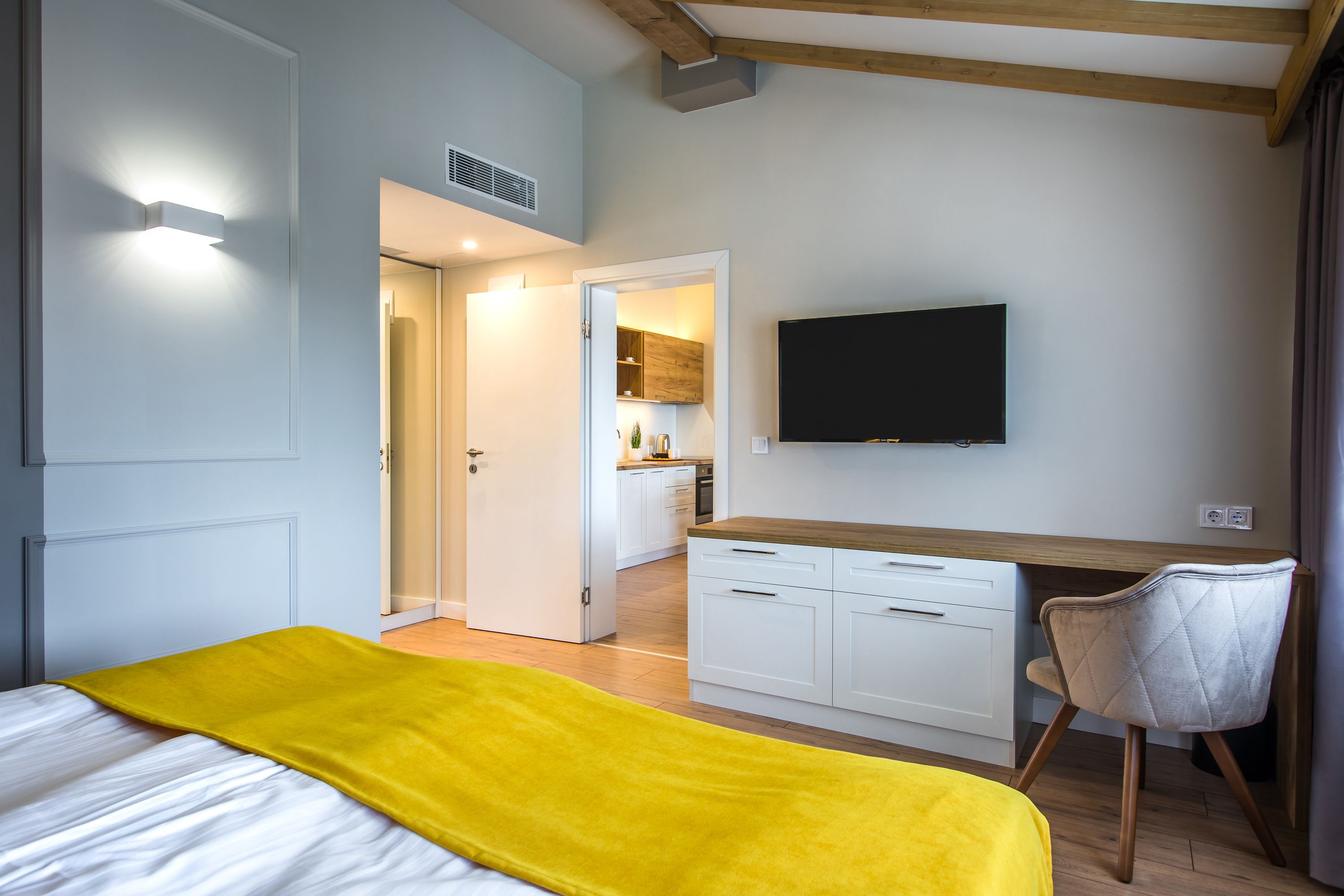Un dormitorio con cama, escritorio y silla. Hay un televisor montado en la pared y una puerta que da a la cocina.