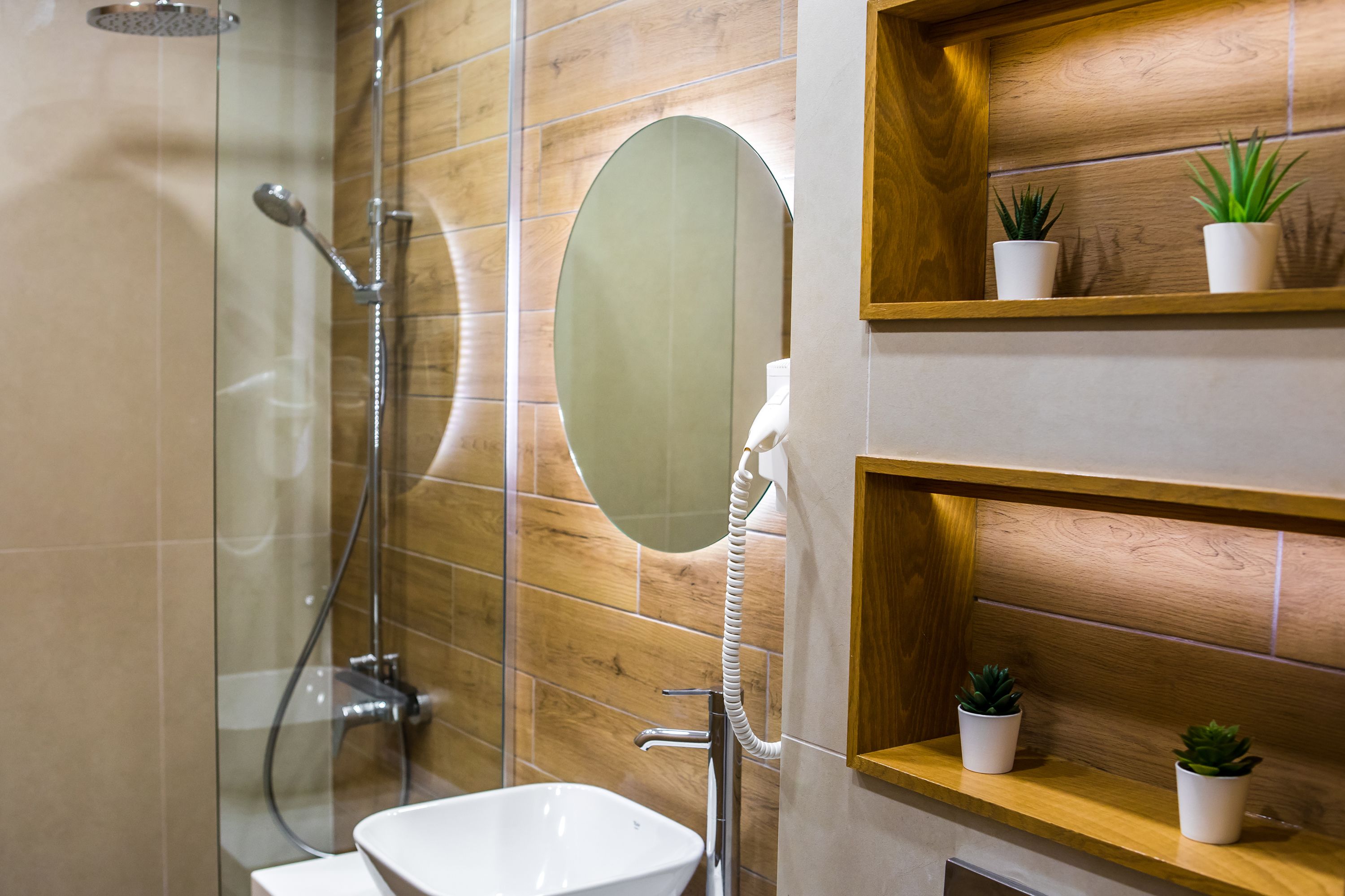 A modern bathroom with a glass-enclosed shower, a white sink, a wooden vanity with two potted plants, and a mirror.