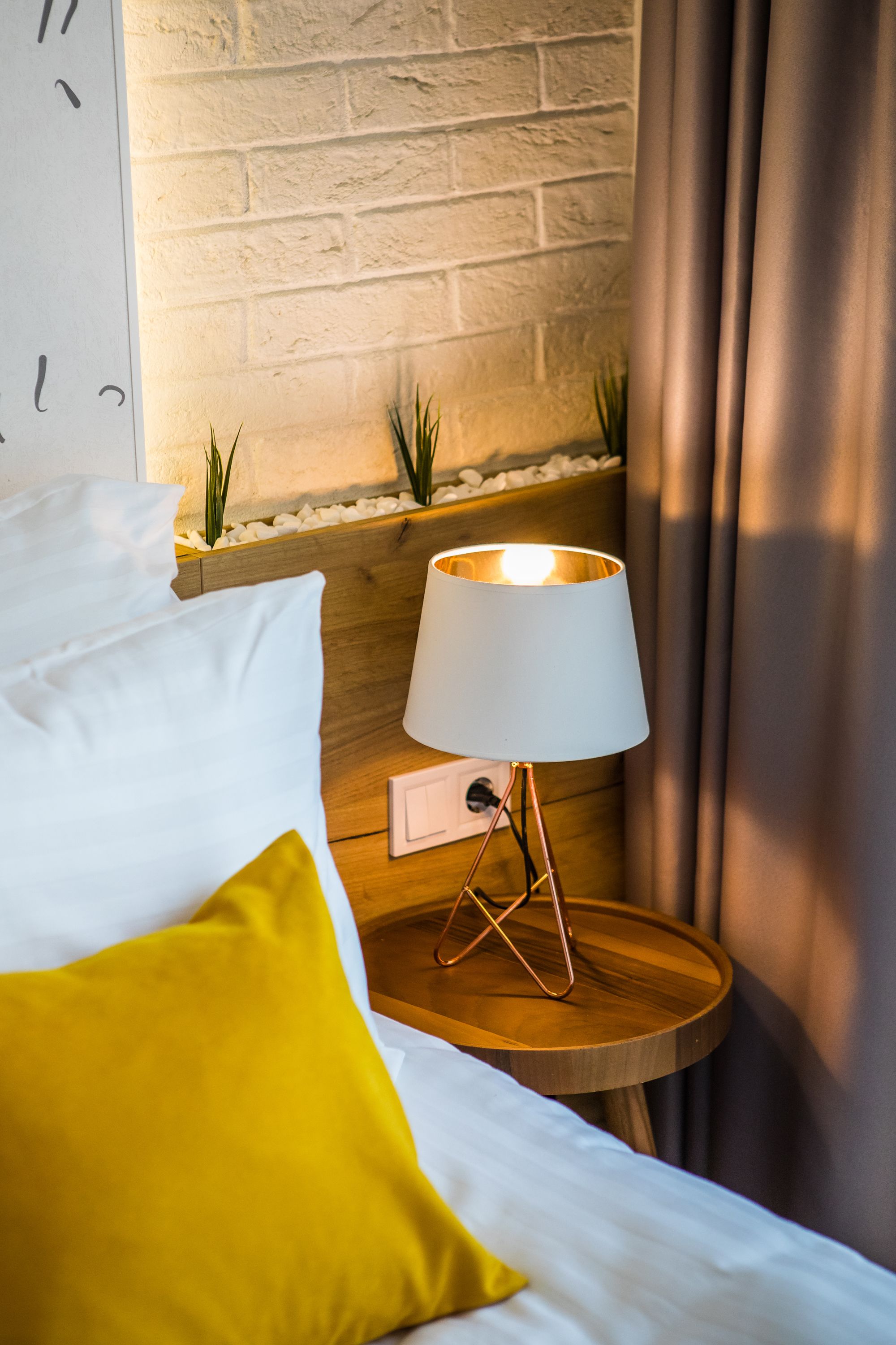 Una cama bien hecha con almohadas blancas y amarillas, una lámpara de noche sobre una mesita de madera y un muro de ladrillo al fondo.
