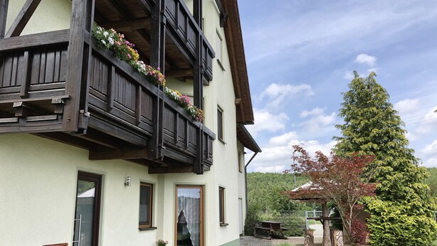 A traditional chalet with wooden balconies, a paved entrance, and a garden with a large tree.