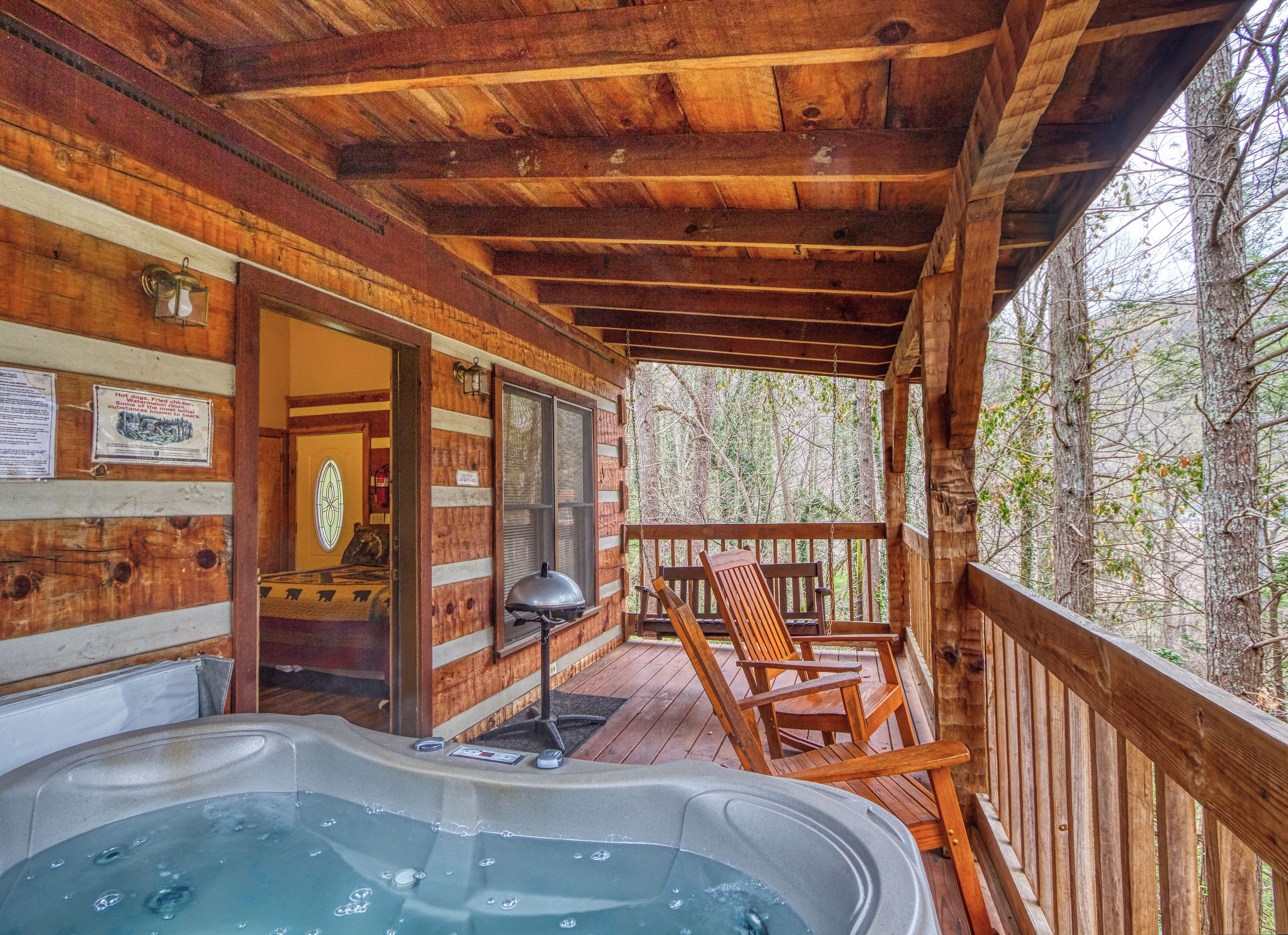 A wooden cabin with a hot tub, wooden deck, and a view of the forest.