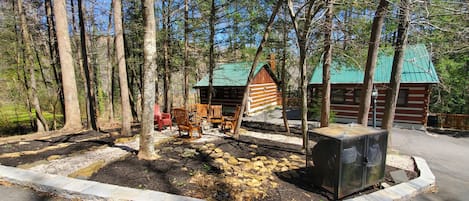 Een rustieke hut met een groen dak, omringd door hoge bomen en een verhard gebied met houten stoelen en bankjes.