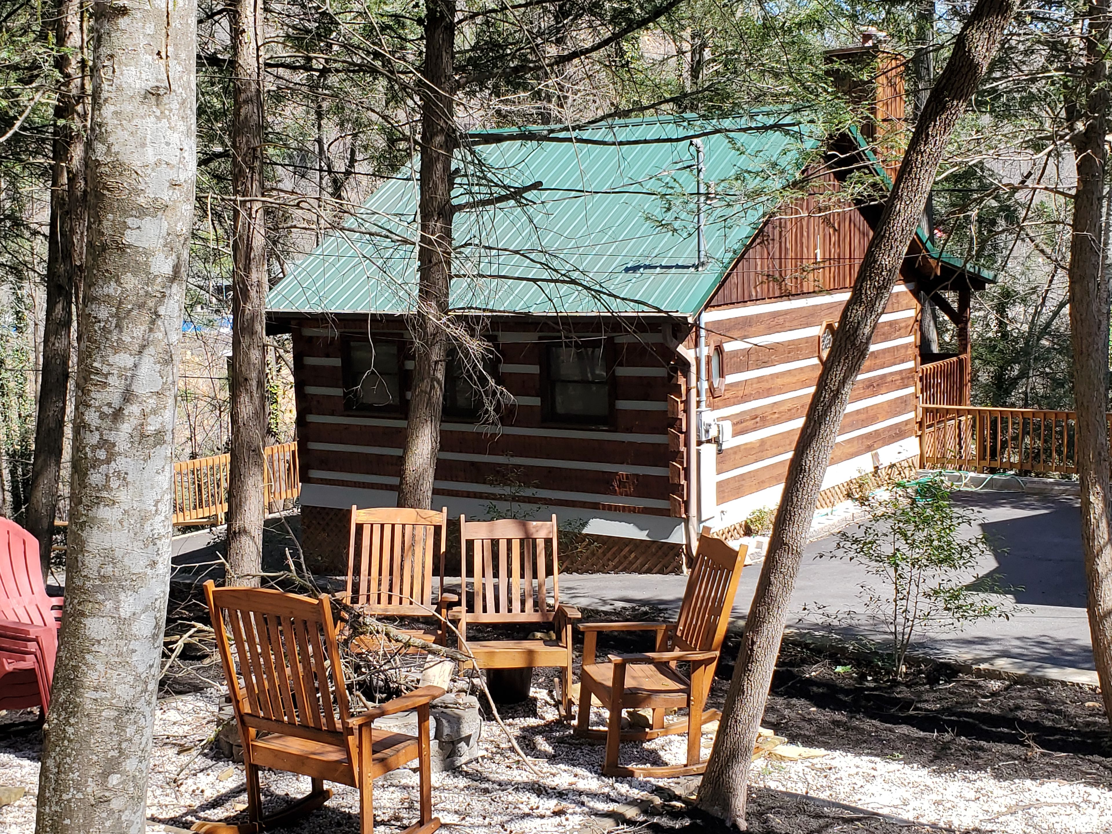 Une cabane rustique avec un toit végétalisé, entourée d’arbres. À l’intérieur, on trouve une table en bois avec des chaises et un foyer.