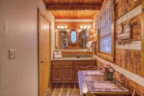 Baño de madera con tocador, lavamanos y espejo. Se ve una ventana con persianas y un adorno en la pared.