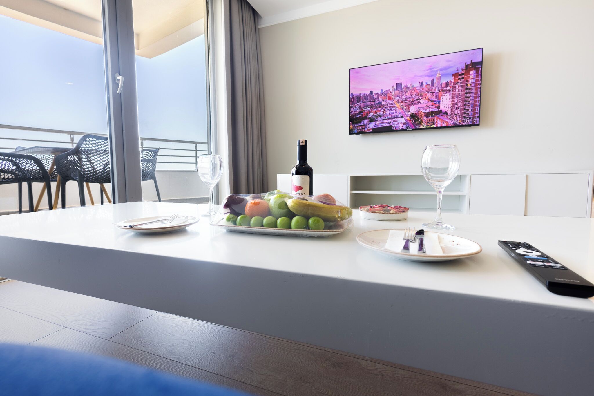 A hotel room with a dining table set with fruits, a bottle of wine, and a remote control.