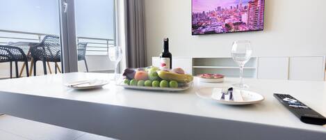 A hotel room with a dining table set with fruits, a bottle of wine, and a remote control.
