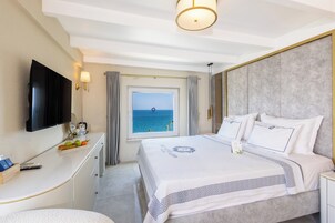 Quarto de hotel com uma cama, escrivaninha, televisão, janela com vista para o mar e uma luminária de teto.