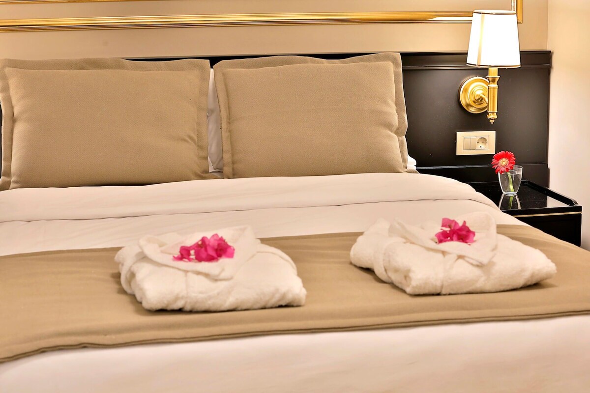 A hotel room with a bed, two pillows, a nightstand with a lamp, and two towels with flowers.