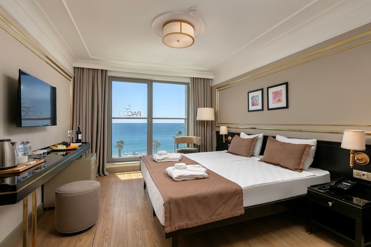 A hotel room with a bed, bedside tables, a desk with a chair, a TV, a view of the ocean, and a window with curtains.