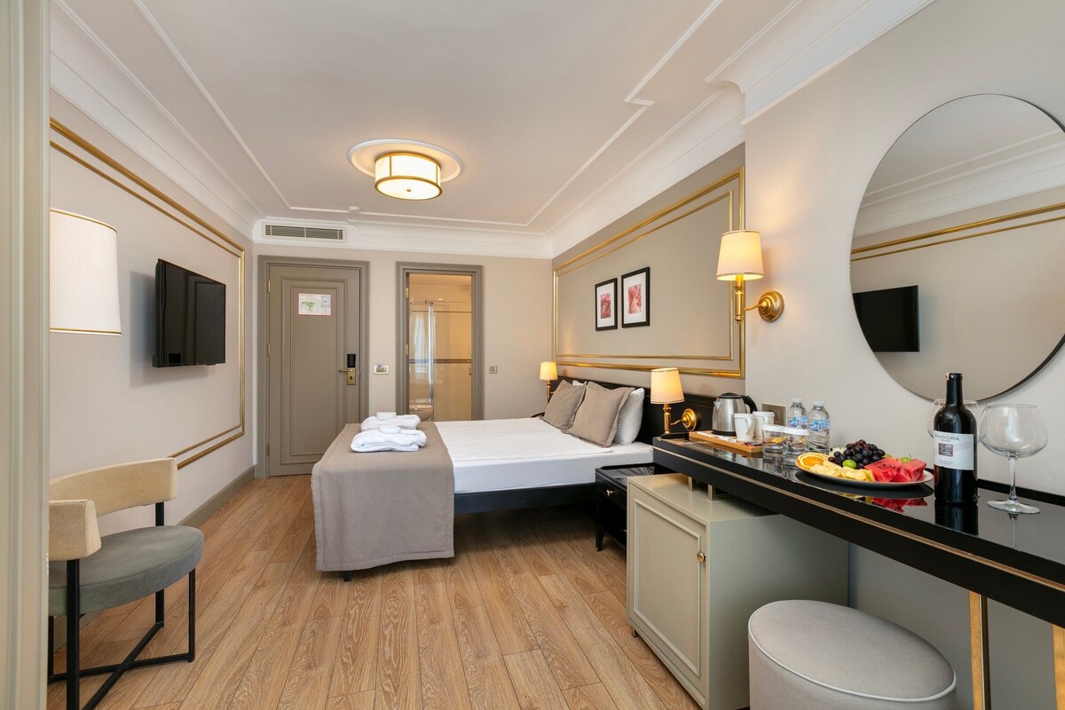 A hotel room with a bed, a small desk with a mirror, a TV, and a minibar.